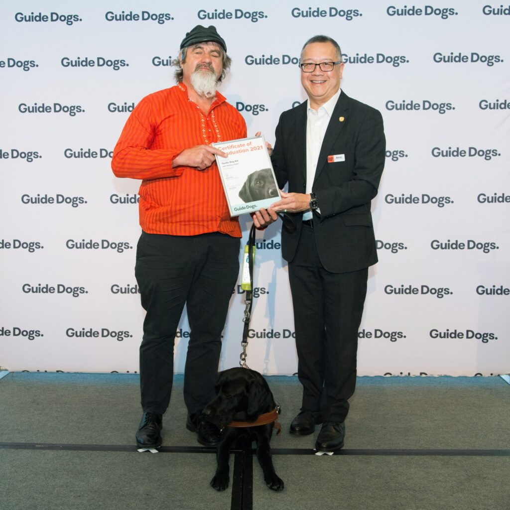 Client Anthony and Guide Dogs SA/NT CEO Aaron Chia standing on stage holding a certificate in front of a Guide Dogs banner. There is a Black Labrador in harness, Kit, sitting between them.