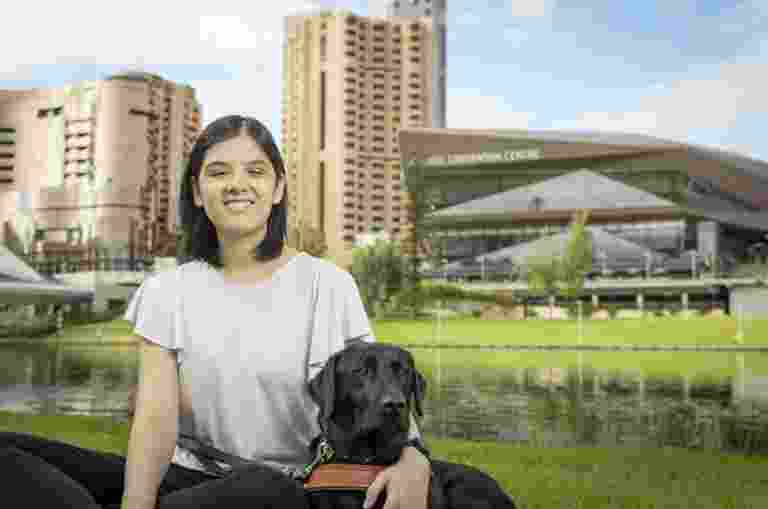 A young woman wearing a white tshirt and black pants is smiling at the camera. Her arm is around a black labrador dog wearing a Guide Dogs harness.