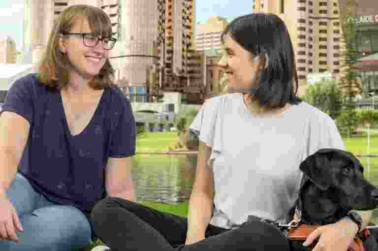Image showing a smiling woman and a girl sitting on green grass in front of the water and Adelaide Convention Centre, they are engaging in eye contact with each other. The girl has her arms around a black labrador guide dog.