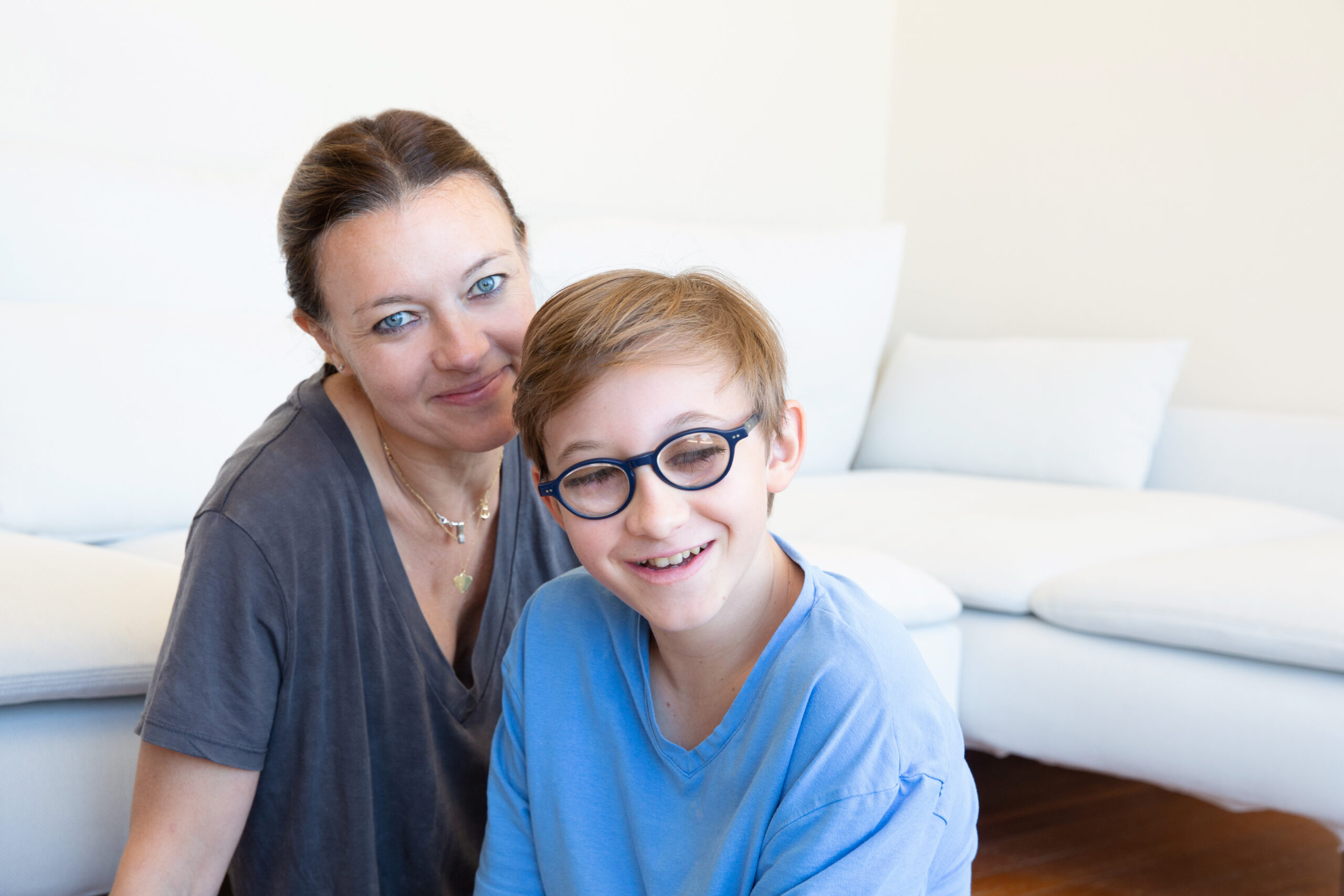 A woman and a young boy with glasses are sitting together in a bright living room, both smiling at the camera.