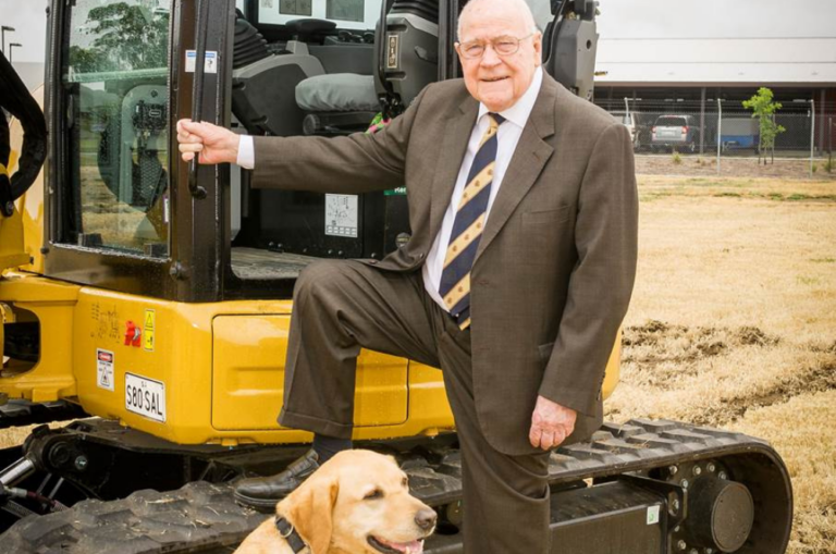 Image shows Frank Beauchamp, Inaugural President of Guide Dogs SA/NT, standing with one foot up on an earthmover in an empty paddock at the sod turning of Beau’s Pet Hotel in 2016. A yellow Labrador, former Ambassadog Tango, sits at his feet wearing a Guide Dogs coat.