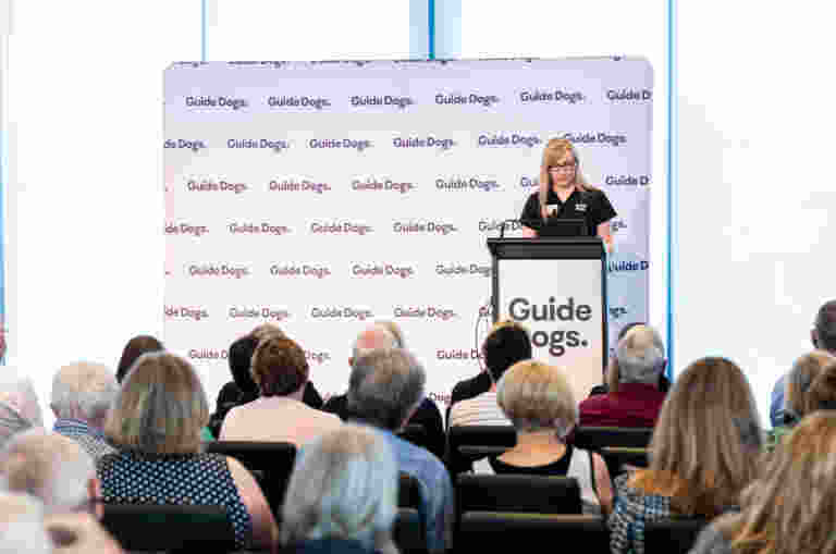 A woman speaks at a podium with Guide Dogs written on it, in front of an audience. A backdrop also displays the words Guide Dogs.