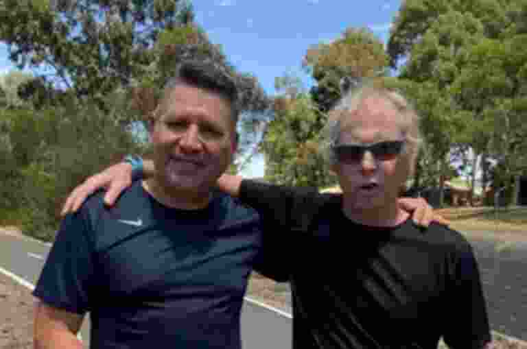 Two men in athletic wear stand side by side on a paved path, one with an arm around the other. Trees and a blue sky are in the background.