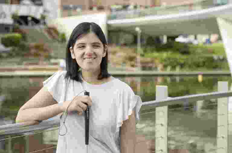 A girl standing against a fence in front of the river torrens holding a white cane.