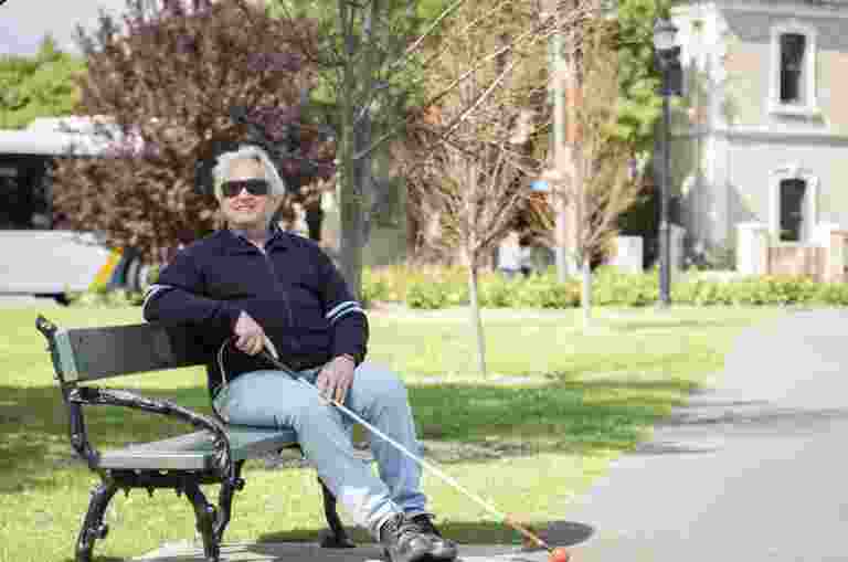 A man with a white cane, sitting on a park bench.
