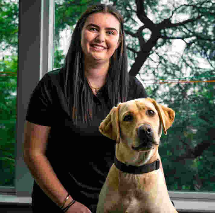 A woman with long dark hair smiles while sitting with a Labrador retriever in front of a window showing green trees.