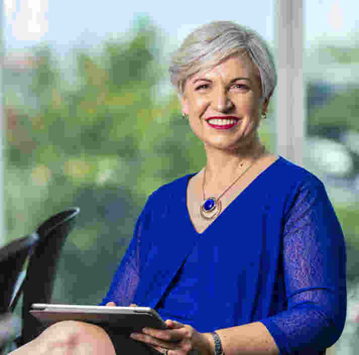 A person with short gray hair wearing a blue outfit is sitting and smiling, holding a tablet.