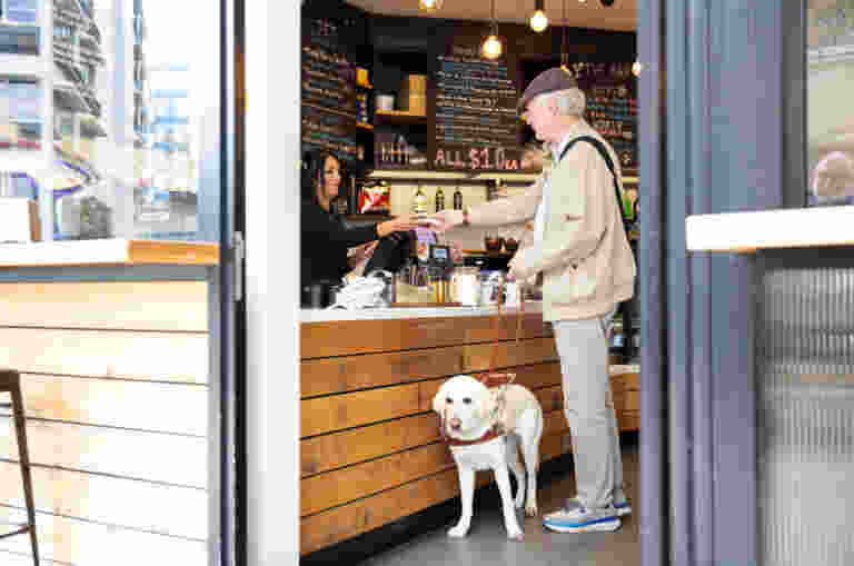 A man with a guide dog pays at a cafe counter, handing money to a barista behind the register.