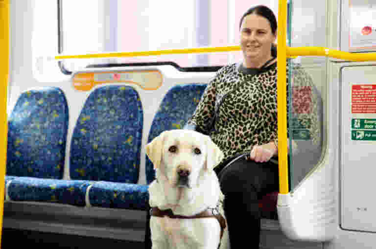 A woman sits on a subway train seat, and a guide dog stands beside her.