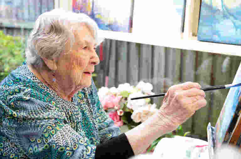An older woman painting a picture in a courtyard garden.