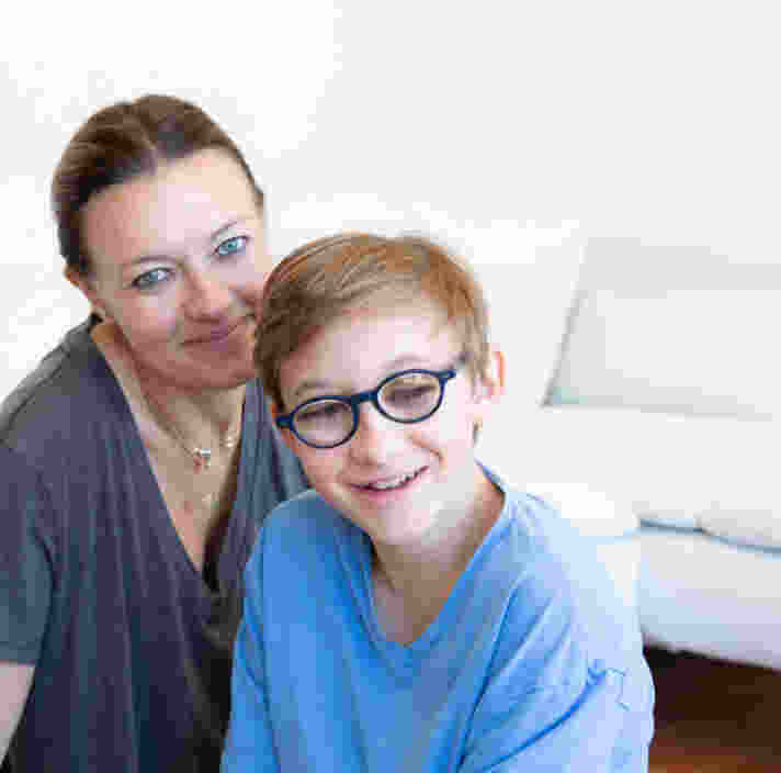 A woman and a child with glasses are sitting indoors, smiling at the camera in front of a white couch.