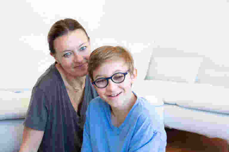 A woman and a child with glasses are sitting indoors, smiling at the camera in front of a white couch.