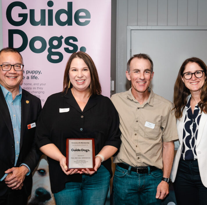 Four people standing in front of a pink banner with the Guide Dogs logo. The people are smiling at the camera and one is holding a honor plaque.