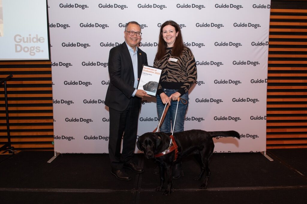 A photo of Claire Stevens and Guide Dog Gwen on stage, happily receiving her certificate.