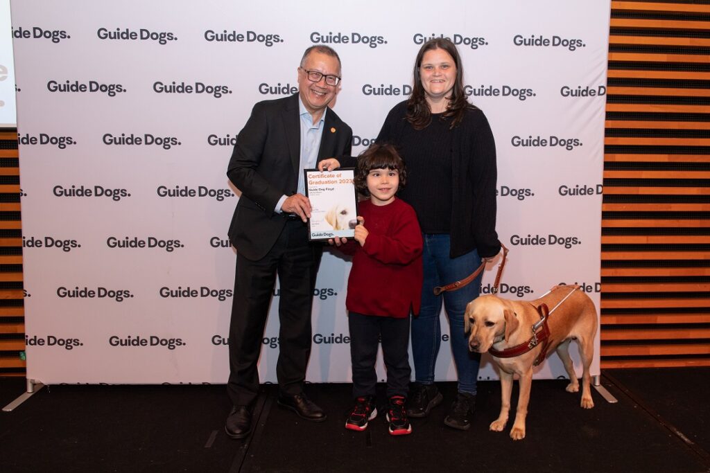 A photo of Lisa Pearce, and her son Leo, and Guide Dog Floyd on stage, happily receiving his certificate.