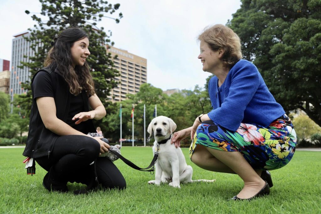 Puppy Training Advisor Cecilia and Her Excellency the Honourable Frances Adamson AC, Governor of South Australia, kneeling on grass with a yellow Labrador puppy.