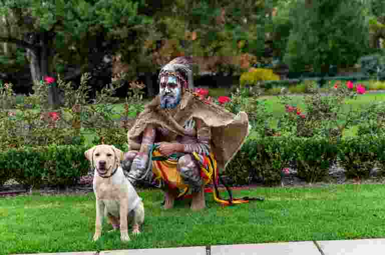 Cliffy Wilson, wearing traditional Aboriginal dress, kneeling on grass with Jarby, a yellow puppy-in-training.