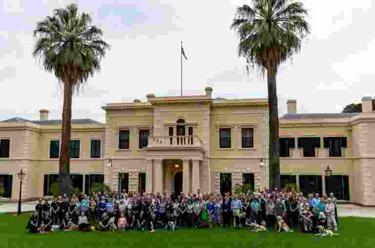 A large group of people and Guide Dogs standing in between two tall palm trees in fron of Government House.