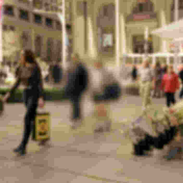 A city street scene with people walking and standing near a flower stand. A person on the left is holding a shopping bag. Background buildings and shops are slightly blurred.