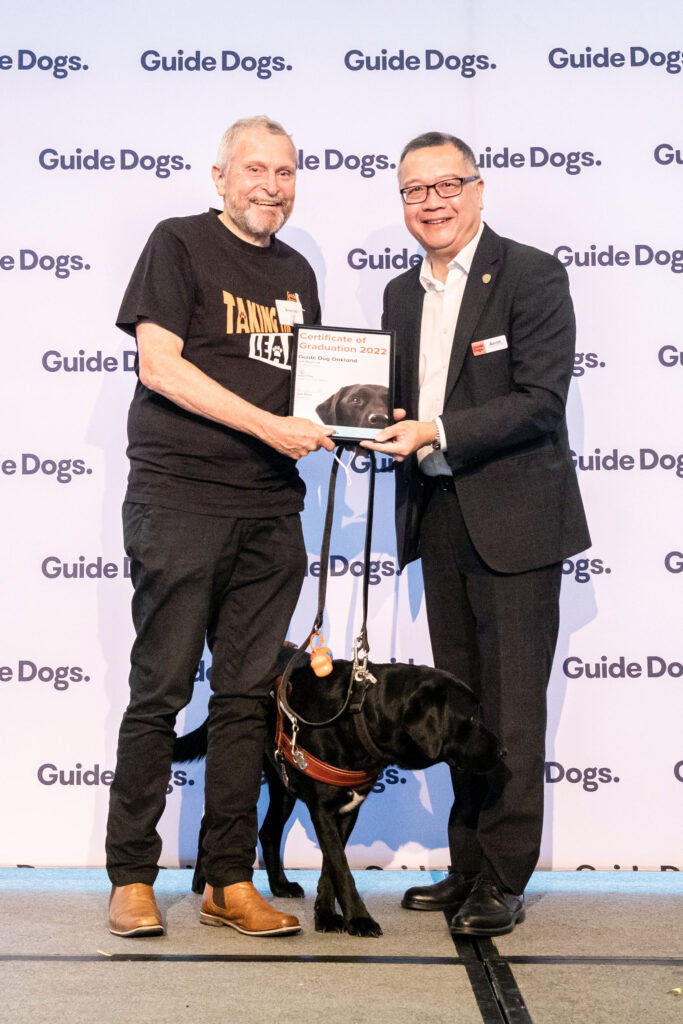 Bruce Ind and Guide Dog Oakland receiving a certificate on stage with Aaron Chia.