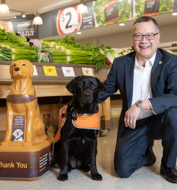 A man in a suit kneels next to a black guide dog wearing an orange harness inside a grocery store, with fresh produce displayed in the background.