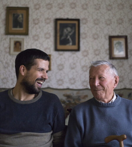 A younger man and an older man sit together on a couch, smiling and looking at each other, with portraits hanging on the wall behind them.