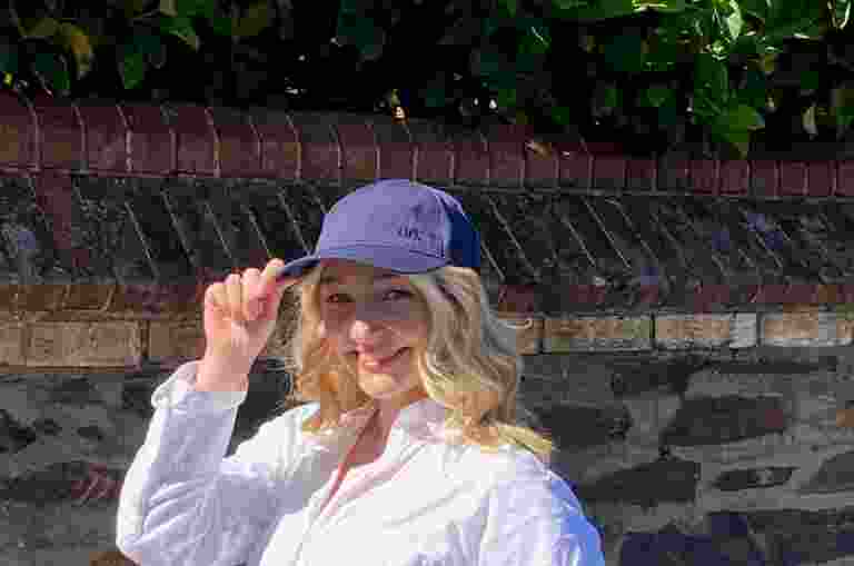 A person in a white shirt and blue cap stands smiling in front of a brick and stone wall, with green foliage above, carrying a Jute Tote Bag.