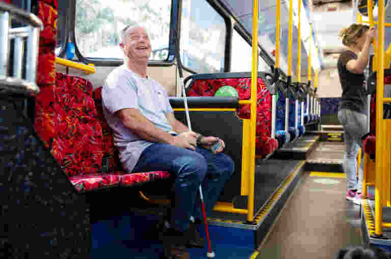 Man holding a white cane smiles while sitting on a bus with red patterned seats.