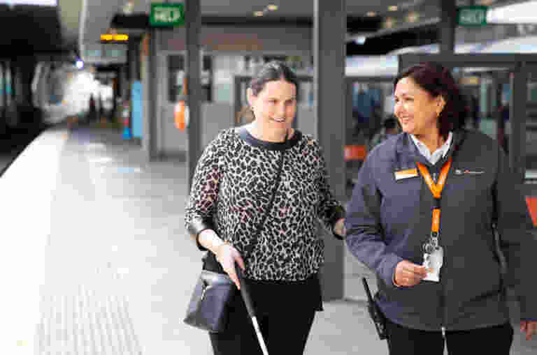 A blind woman with a cane is guided by a transit staff member at a train station platform, with trains and signage visible in the background.