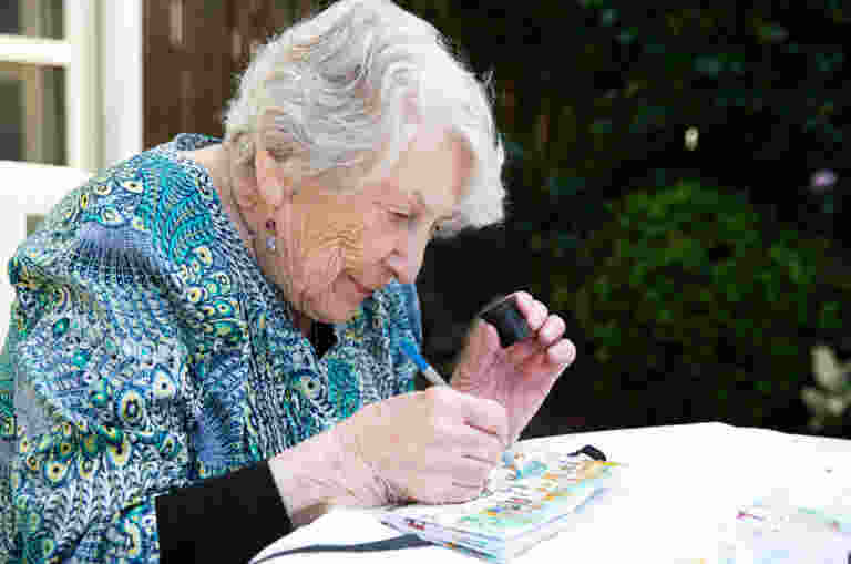 An elderly woman with white hair is sitting outdoors, painting with watercolors on paper, focused intently.