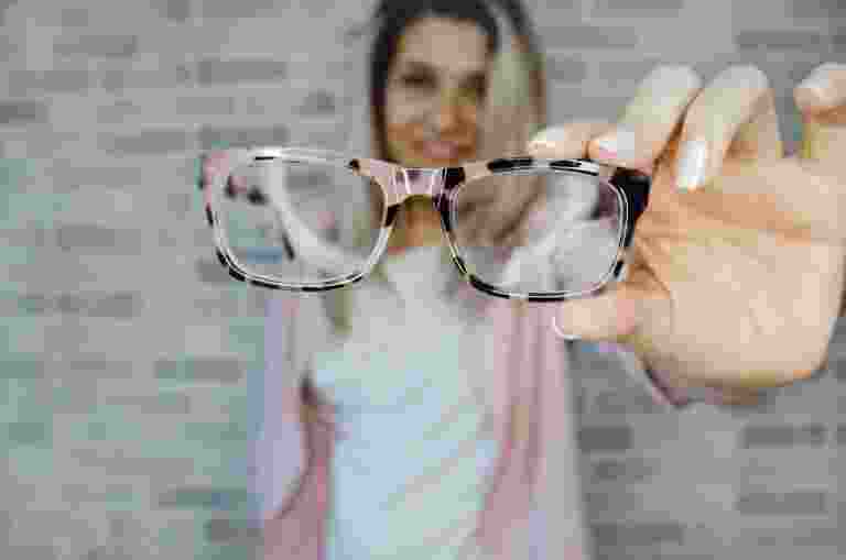 A person holds a pair of eyeglasses toward the camera with a blurred brick wall background.