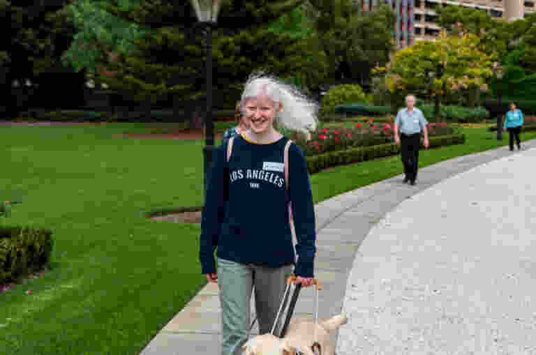 A person with white hair walks a golden retriever on a sidewalk in a park. A tall building is visible in the background.