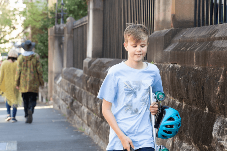A boy carrying a skateboard and helmet walks along a sidewalk with a cane, next to a stone wall.
