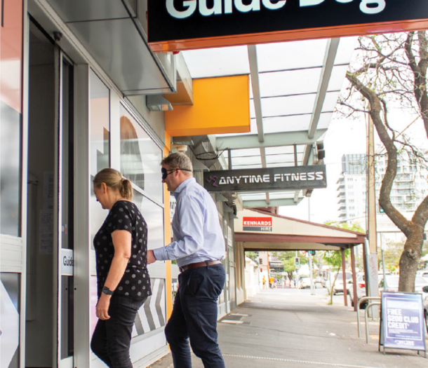 A man and woman entering a building that has a Guide Dogs sign at the entrance.