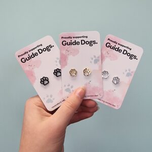 Image shows a hand holding three pairs of paw-shaped stud earrings, each displayed on a card that says "Proudly supporting Guide Dogs." The left pair is black coloured, the middle pair is gold and the pair on the right is silver coloured. The background is a plain light blue wall.