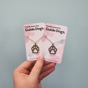 Image shows a hand holding two necklaces with paw-shaped pendants, each displayed on a card that says "Proudly supporting Guide Dogs." The left pendant is gold-colored with a matching chain, and the right pendant is silver-colored with a matching chain. The background is a plain light blue wall.