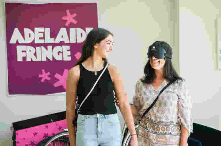 Two women stand smiling, one wearing VR goggles. The background features a sign reading Adelaide Fringe.