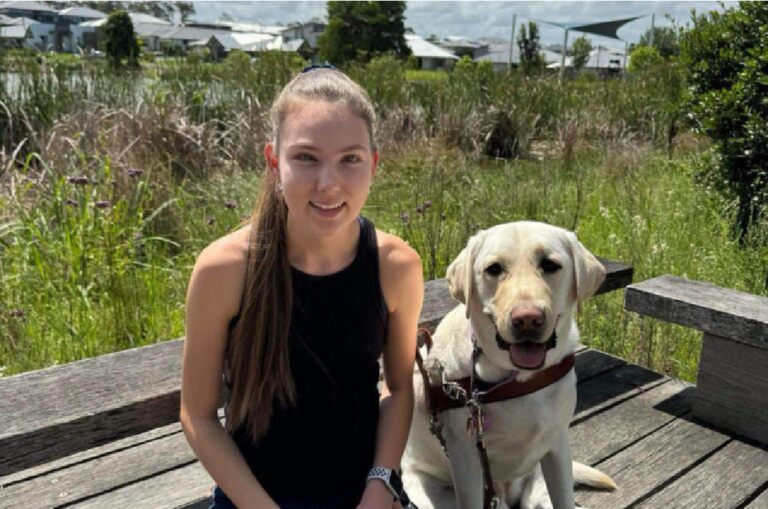 A woman sitting on a bench next to a yellow Labrador retriever guide dog, outdoors in a park setting.