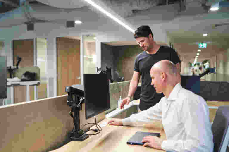 Two men in an office setting, one standing and pointing at a computer screen while the other sits at a desk, observing.