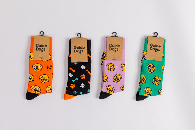 Four pairs of Socks - Coloured, featuring dog and paw print designs, are displayed side by side with Guide Dogs labels on cardboard tags.