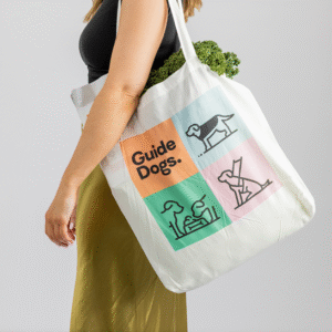 A person carries a white Bag - Cotton with Guide Dogs and colorful guide dog illustrations on it, filled with leafy greens.