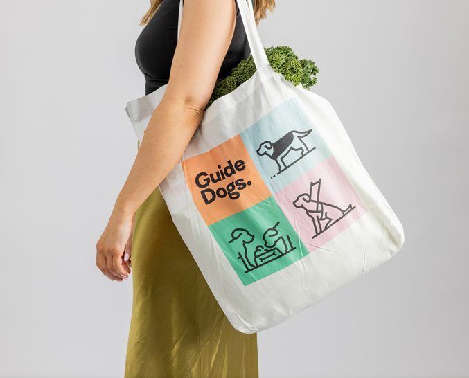 A person carries a white Bag - Cotton with Guide Dogs and colorful guide dog illustrations on it, filled with leafy greens.