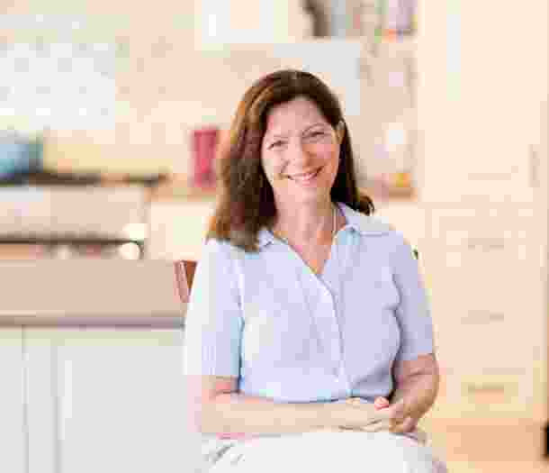 Smiling woman with long brown hair sits in a bright kitchen, wearing a light blue sweater and white pants.