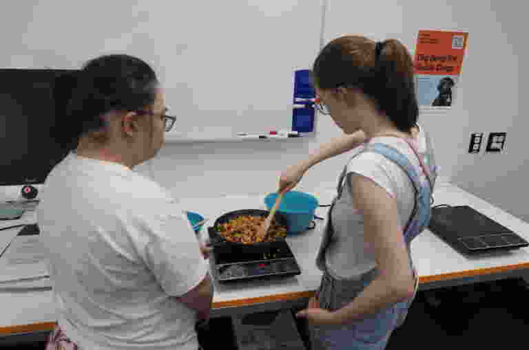 Two clients cooking in a kitchen. One person stirs a pan filled with food on a portable stove.