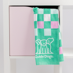 A Tea Towel - Puppy Icons (2 Pack) featuring green, white, and pink checks and a Guide Dogs logo hangs over a white shelving unit with a pink storage cube.