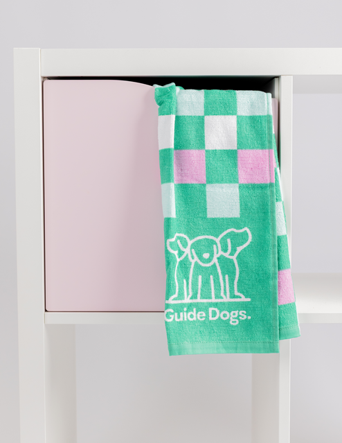 A Tea Towel - Puppy Icons (2 Pack) featuring green, white, and pink checks and a Guide Dogs logo hangs over a white shelving unit with a pink storage cube.