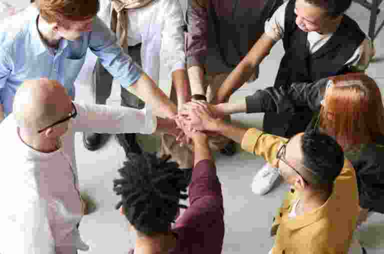A group of people stand in a circle with their hands joined in the center, symbolizing support.
