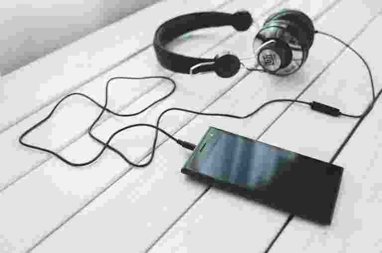 Image portraying black over-ear headphones with a cord connected to a smartphone on a white wooden surface.