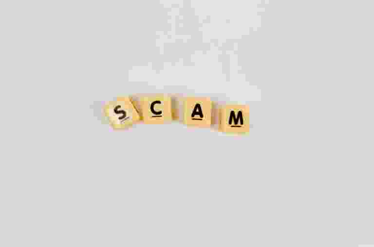 Four square scrabble letter tiles on a white surface spell the word SCAM.