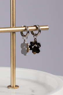 Two Huggie Paws metal hoop earrings, one silver and one black with flower-shaped charms, hang on a horizontal gold rod against a neutral background.